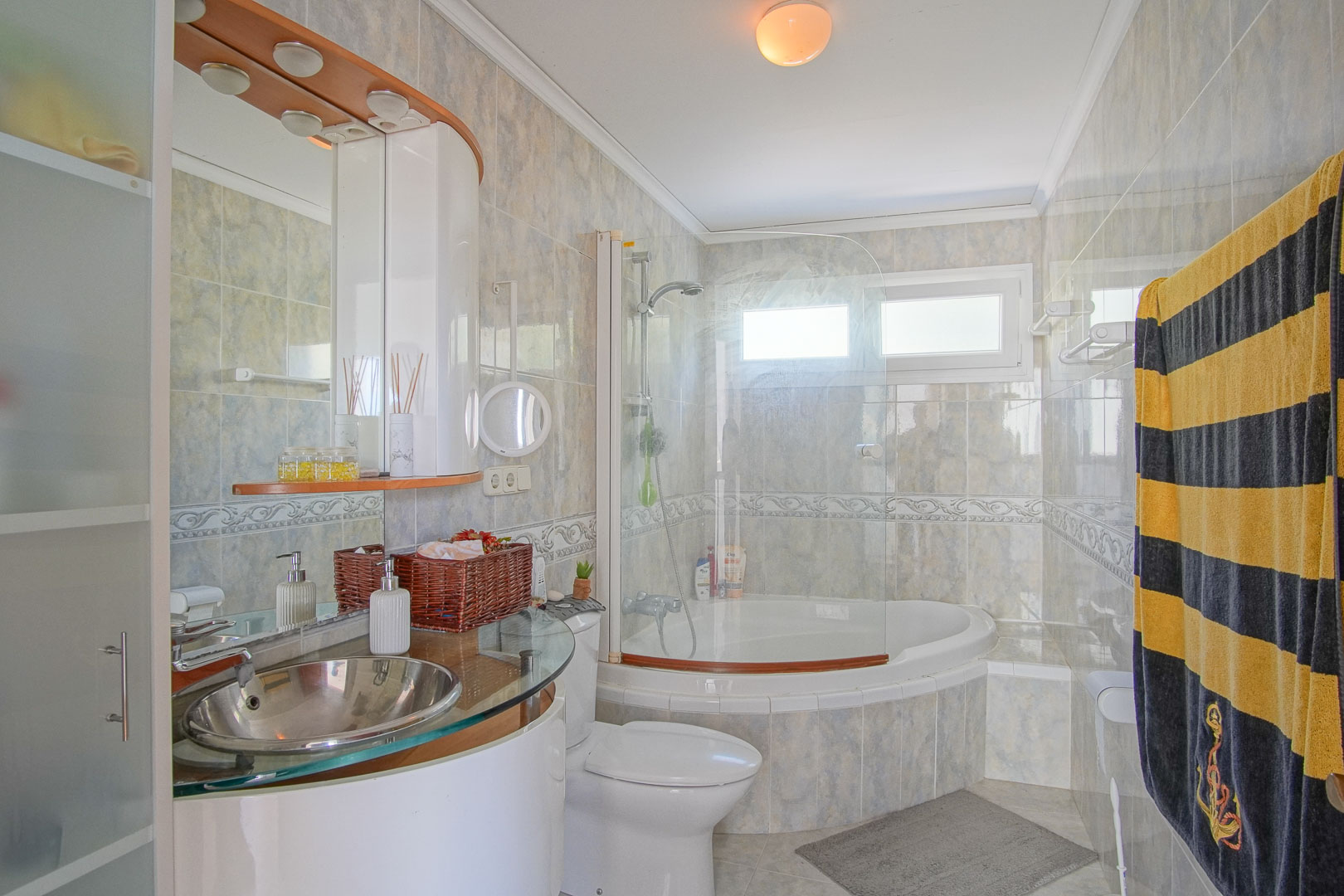 Complete badkamer met hoekbad, glazen douchescherm en moderne roestvrijstalen waskom op een glazen blad. Betegelde wanden met decoratieve rand en natuurlijk licht.