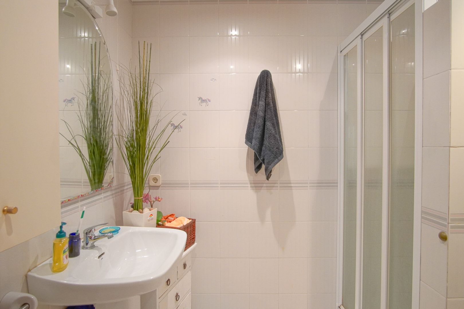 Functionele badkamer met wit betegelde wanden, een porseleinen wastafel, boogspiegel en douchecabine met witte schuifdeur.