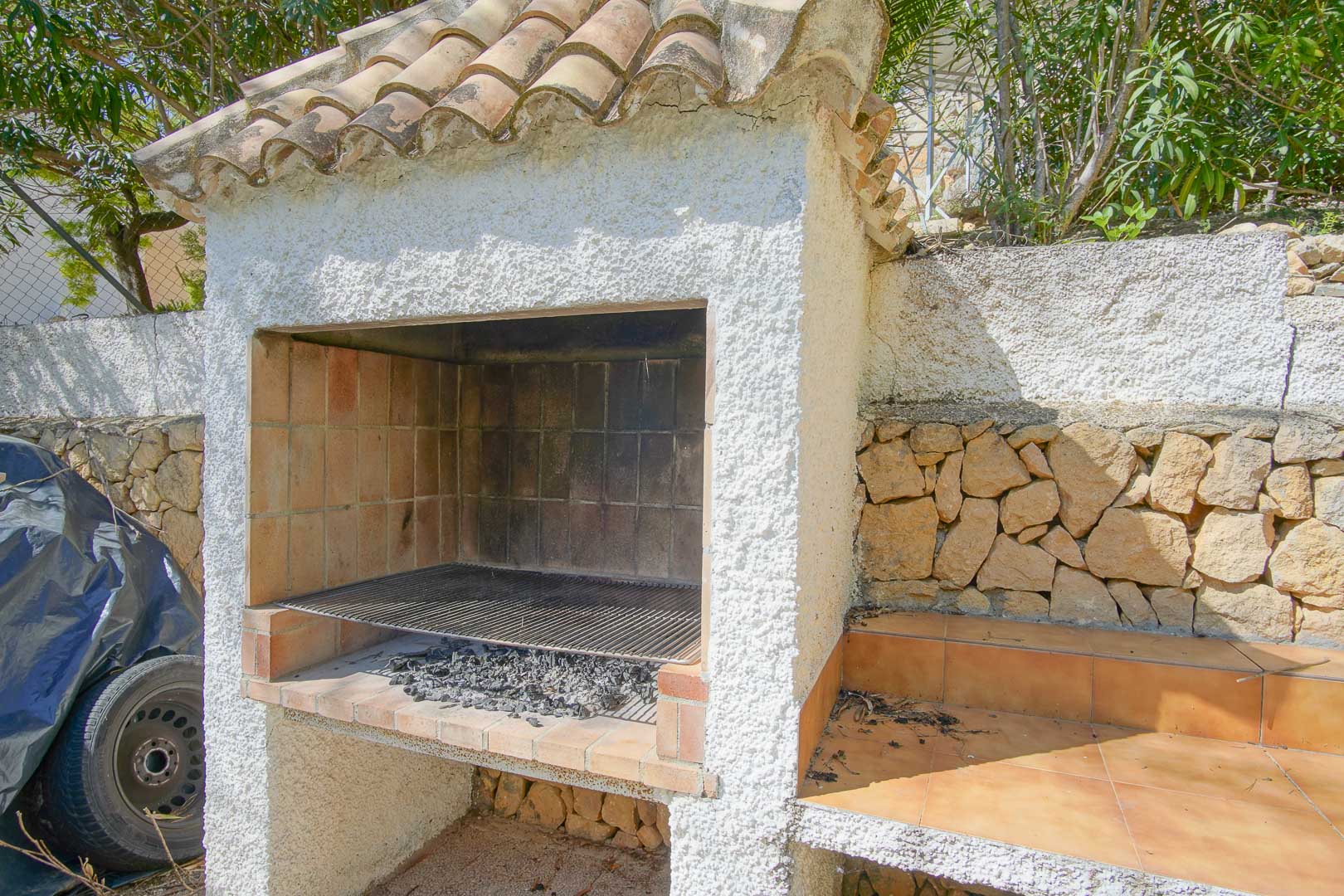 Gemetselde buitenbarbecue met een dak van terracotta pannen en witte stuclaag, geplaatst tegen een natuurstenen muur met een betegeld werkblad.