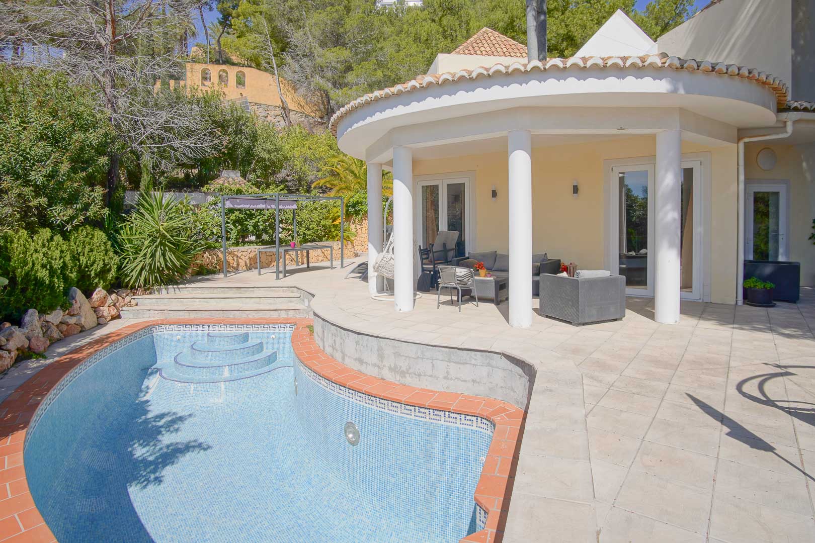 Halfronde overdekte veranda met witte zuilen grenzend aan een privézwembad en zonneterras, gelegen in een volgroeide mediterrane tuin.
