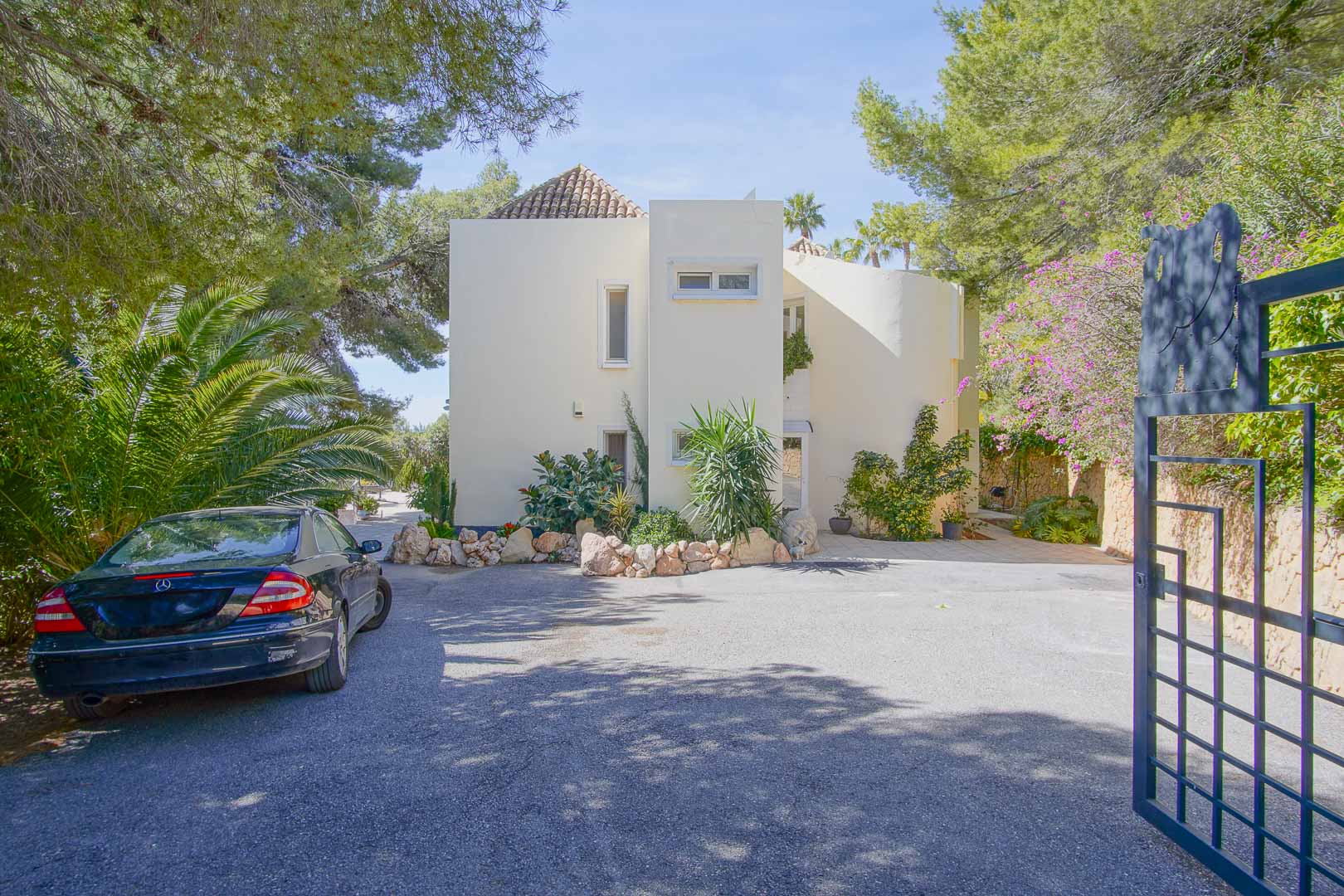 Hoofdfaçade van de villa in mediterrane stijl met een ruime geplaveide parkeerplaats en een beveiligde ingang, omgeven door pijnbomen en weelderig groen.