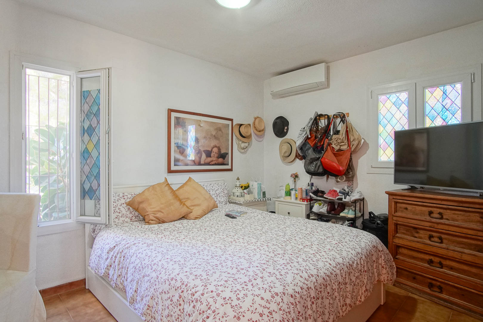 Lichte slaapkamer met tegelvloer, split-airconditioning en ramen met decoratief glas-in-lood. Voorzien van een klassieke houten ladekast en natuurlijke lichtinval.