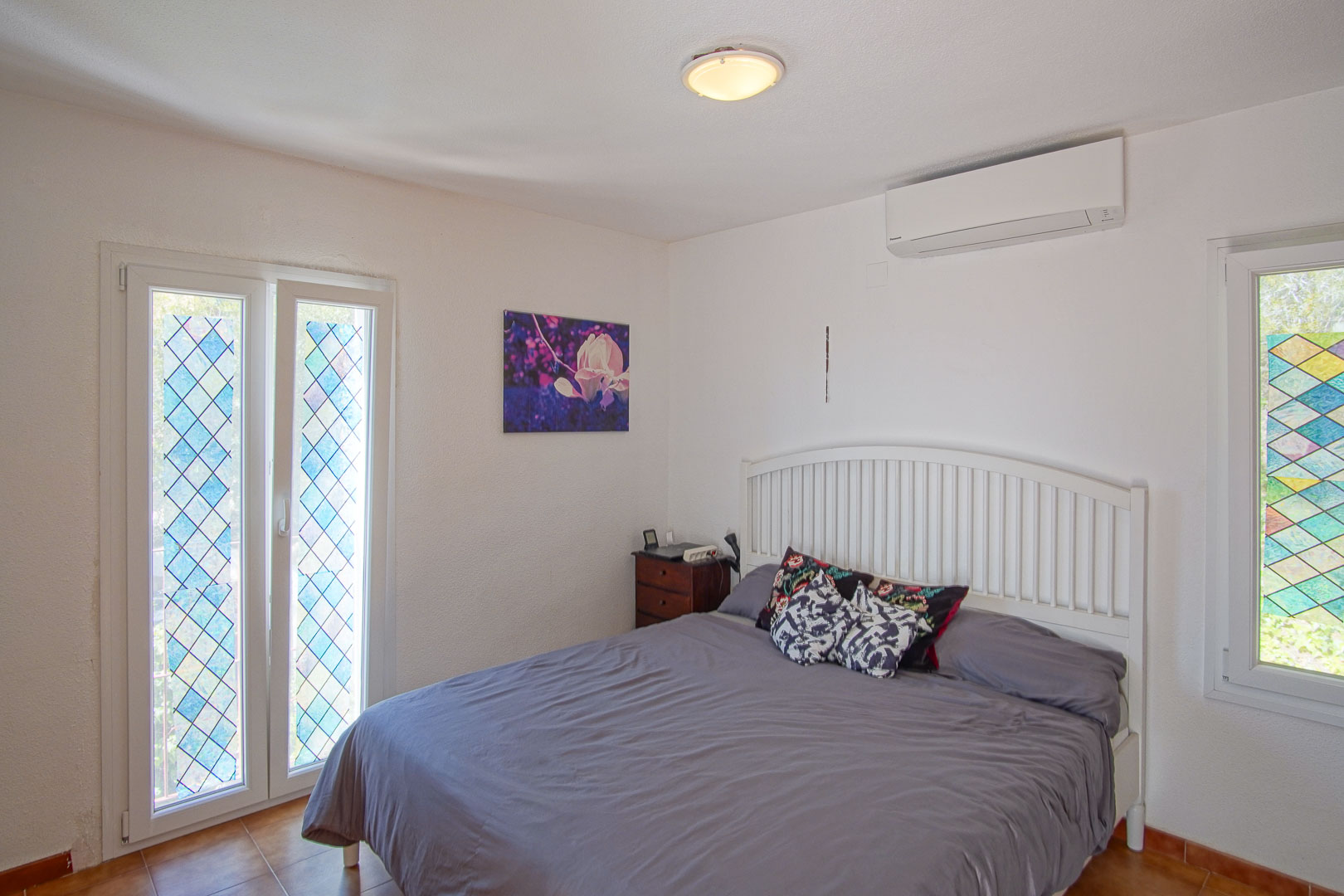 Lichte slaapkamer voorzien van airconditioning, een wit hoofdbord en ramen met decoratief glas-in-lood in ruitpatroon.
