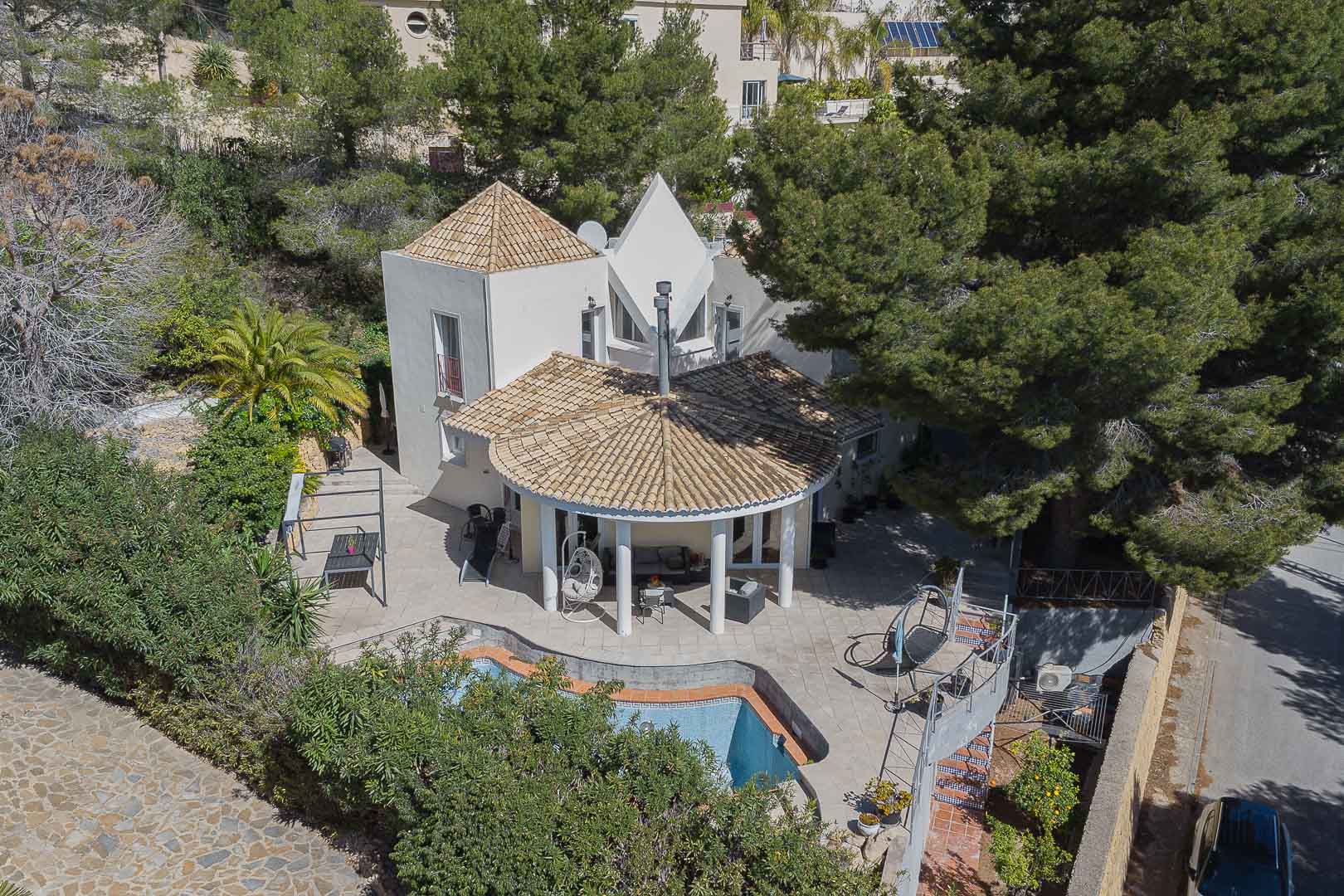 Luchtfoto van de villa met een ronde veranda met witte zuilen, privézwembad en geplaveide terrassen, omgeven door volgroeide pijnbomen en mediterrane begroeiing.