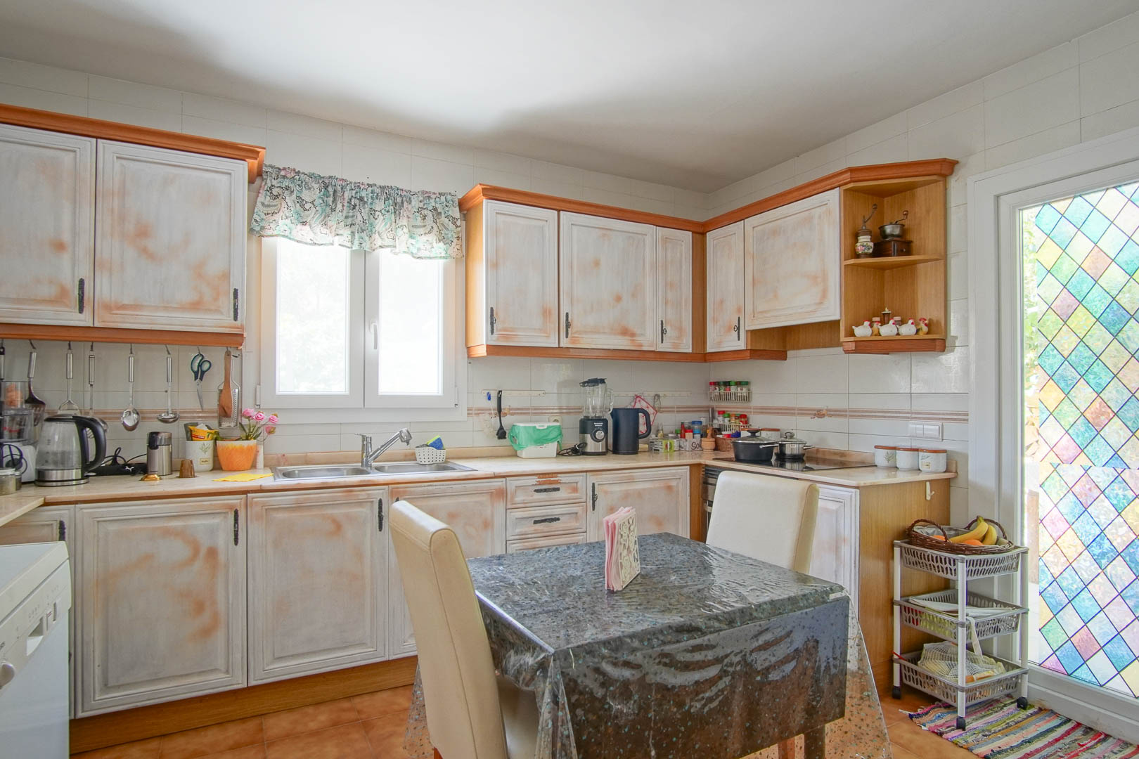 Onafhankelijke rustieke keuken met kasten in verweerde look, een L-vormig werkblad en een eethoek. De ruimte geniet van natuurlijk licht en een decoratieve glas-in-looddeur.