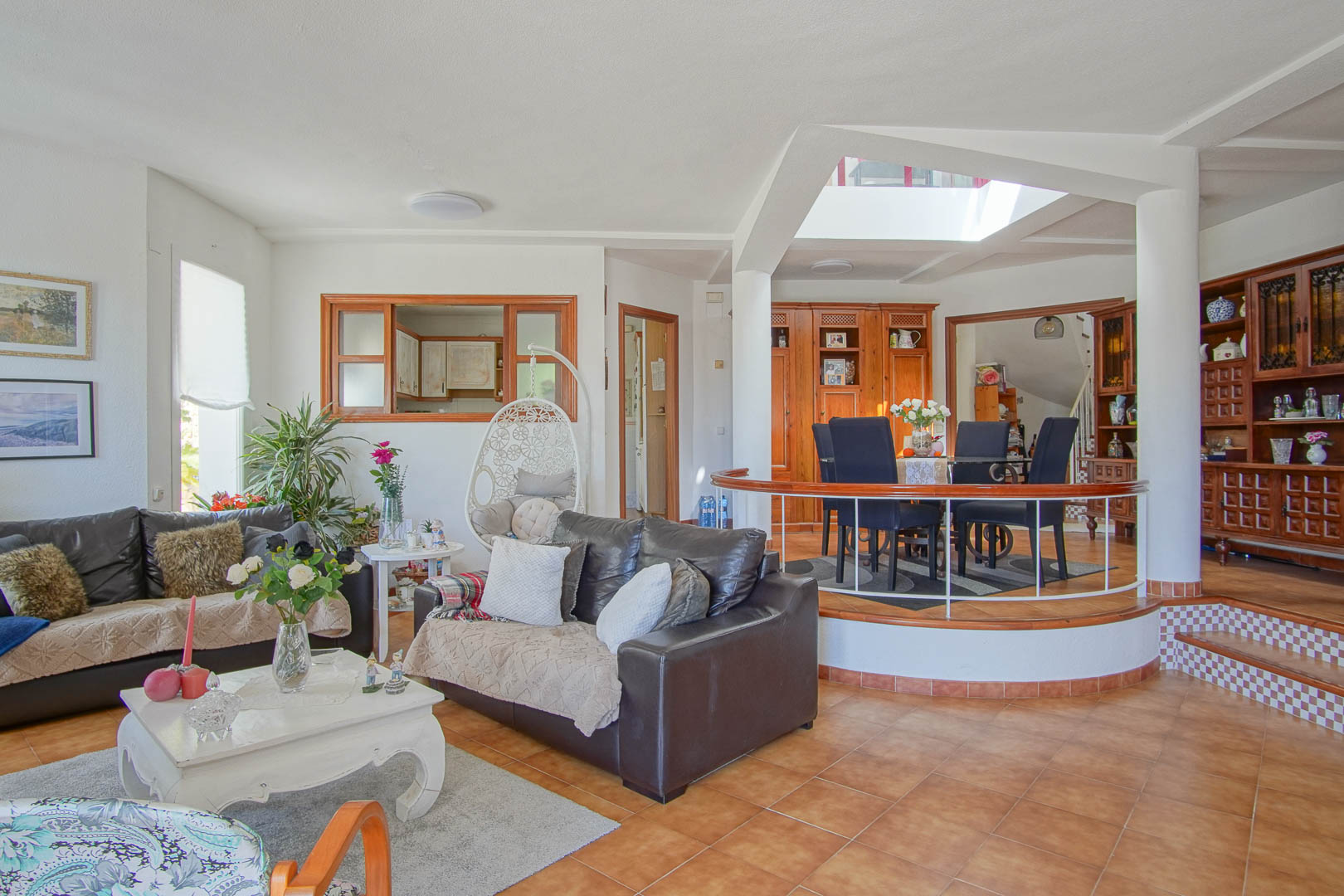 Open woonkamer met terracotta tegels, lederen banken en een verhoogd eetgedeelte op een rond plateau, verlicht door een centraal dakraam voor optimaal daglicht.