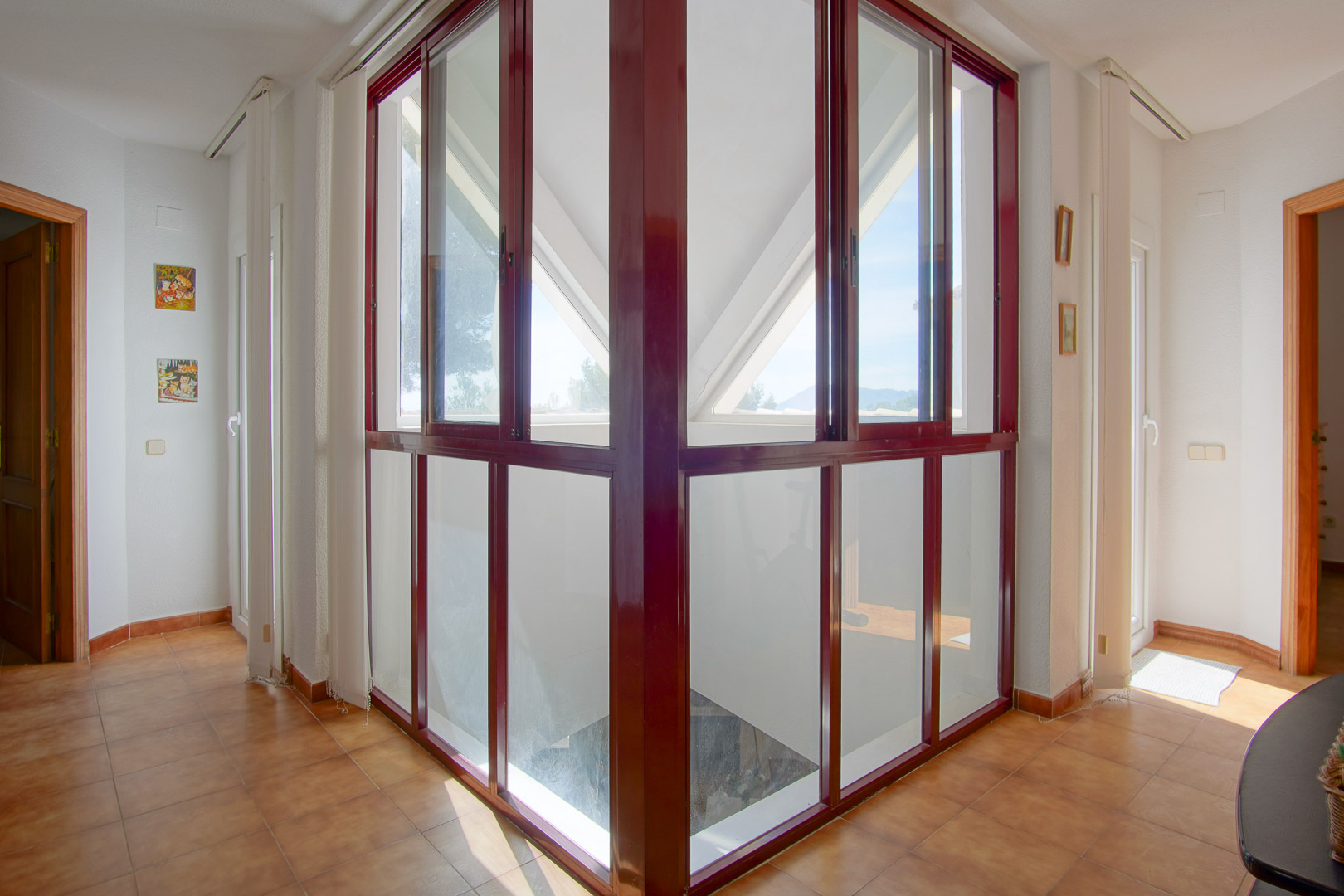 Overloop op de bovenverdieping met een glazen hoekconstructie in bordeauxrode kozijnen, terracotta tegels en veel natuurlijk lichtinval tussen de kamers.