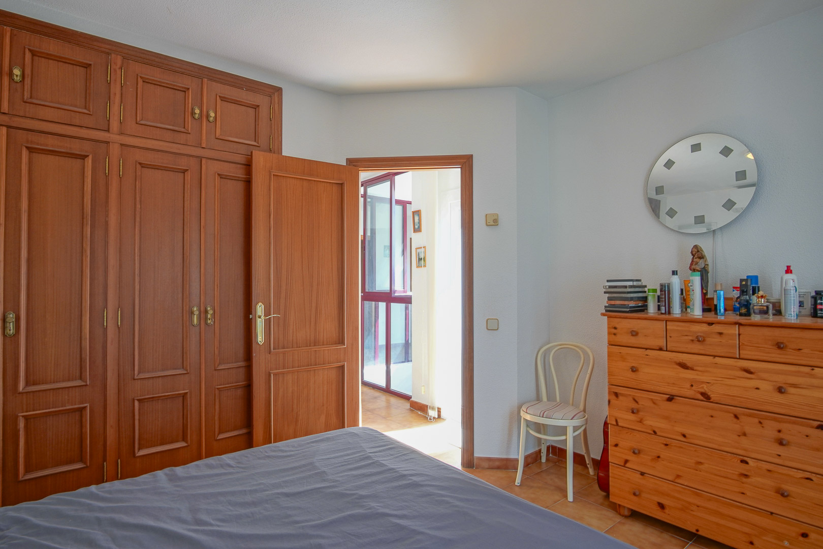 Slaapkamer met kamerhoge houten inbouwkasten, een bijpassende ladekast en een tegelvloer. De kamer staat in directe verbinding met een lichte gang via een klassieke houten deur.
