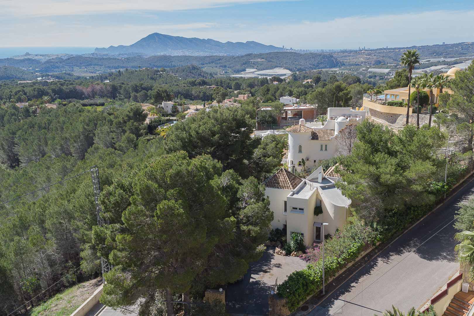 Verhoogd bovenaanzicht van de villa omgeven door pijnbomen, met de mediterrane architectuur en een panoramisch uitzicht op de zee en de bergen.