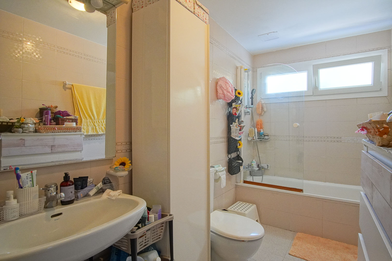 Volledige badkamer met ligbad en glazen scherm, witte wastafel en hoge opbergkast. Beige wandtegels en een venster zorgen voor natuurlijke lichtinval.