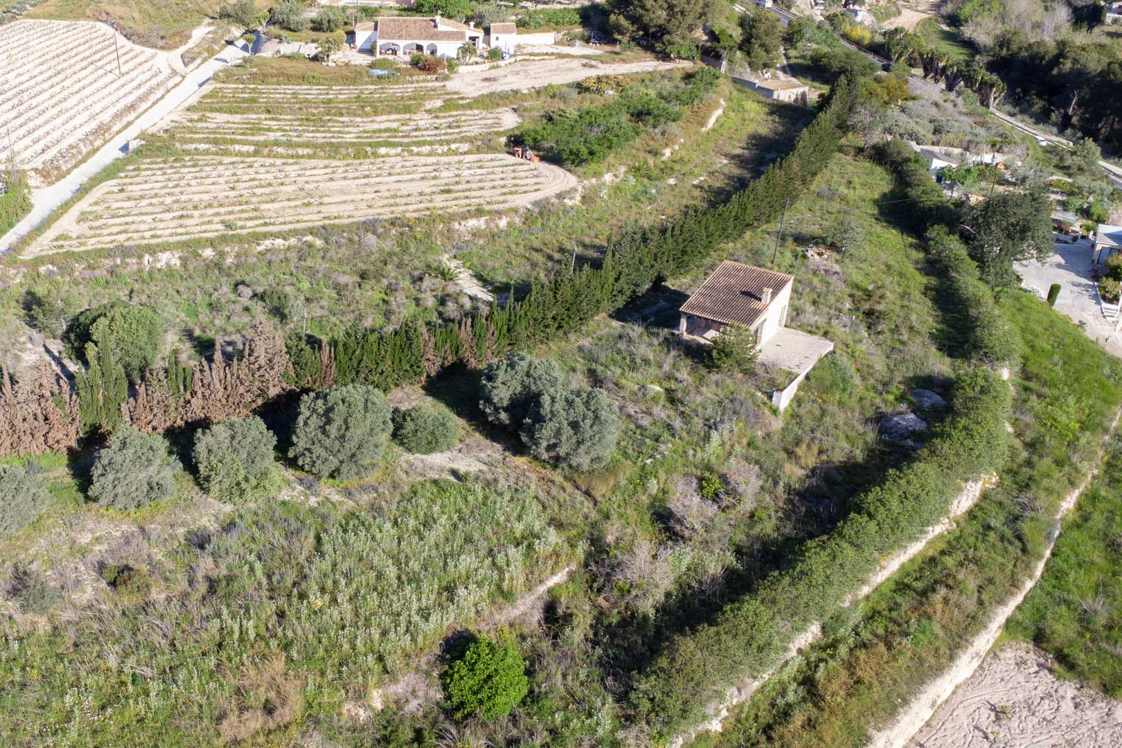 Luchtfoto van het perceel van 10.920 m2 in Teulada, met terrasvormig terrein, mediterrane vegetatie en een bestaand landelijk bijgebouw.