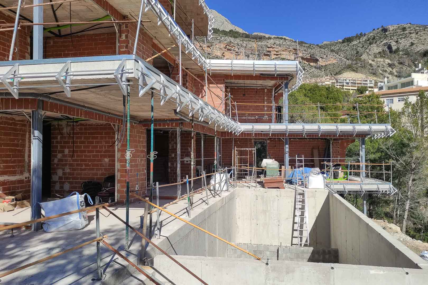 Bouwfase van de villa in Altea, met zicht op de bakstenen muren, betonvloeren en de ruwbouw van het zwembad tegen de berghelling.
