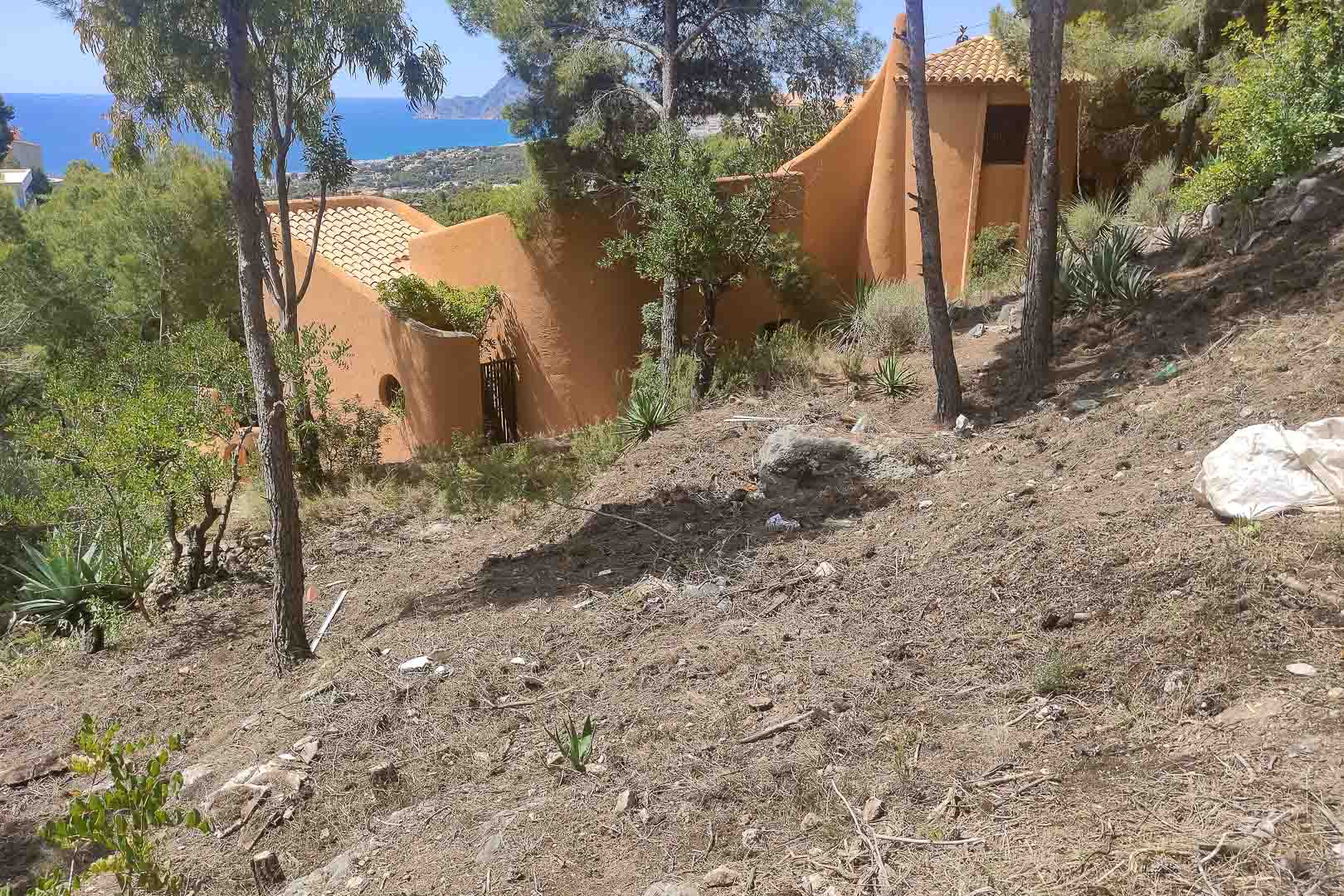 Buitenaanzicht van de villa met organische architectuur en terracotta gevels, gelegen op een hellend perceel met pijnbomen en panoramisch uitzicht op de Middellandse Zee.
