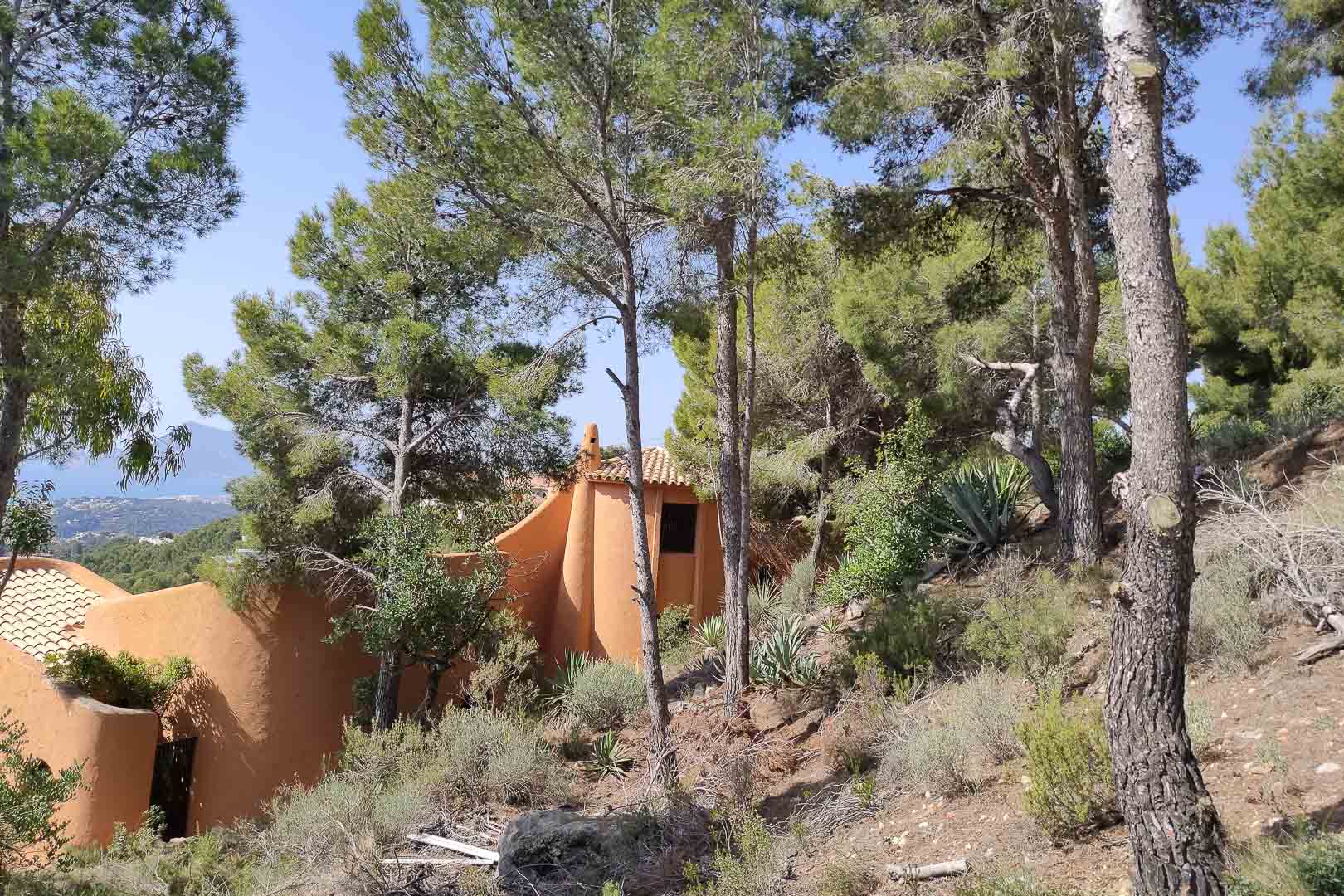 Externe weergave van de organische architectuur in terracottatinten, verscholen in een dicht dennenbos en mediterrane vegetatie met uitzicht op zee.