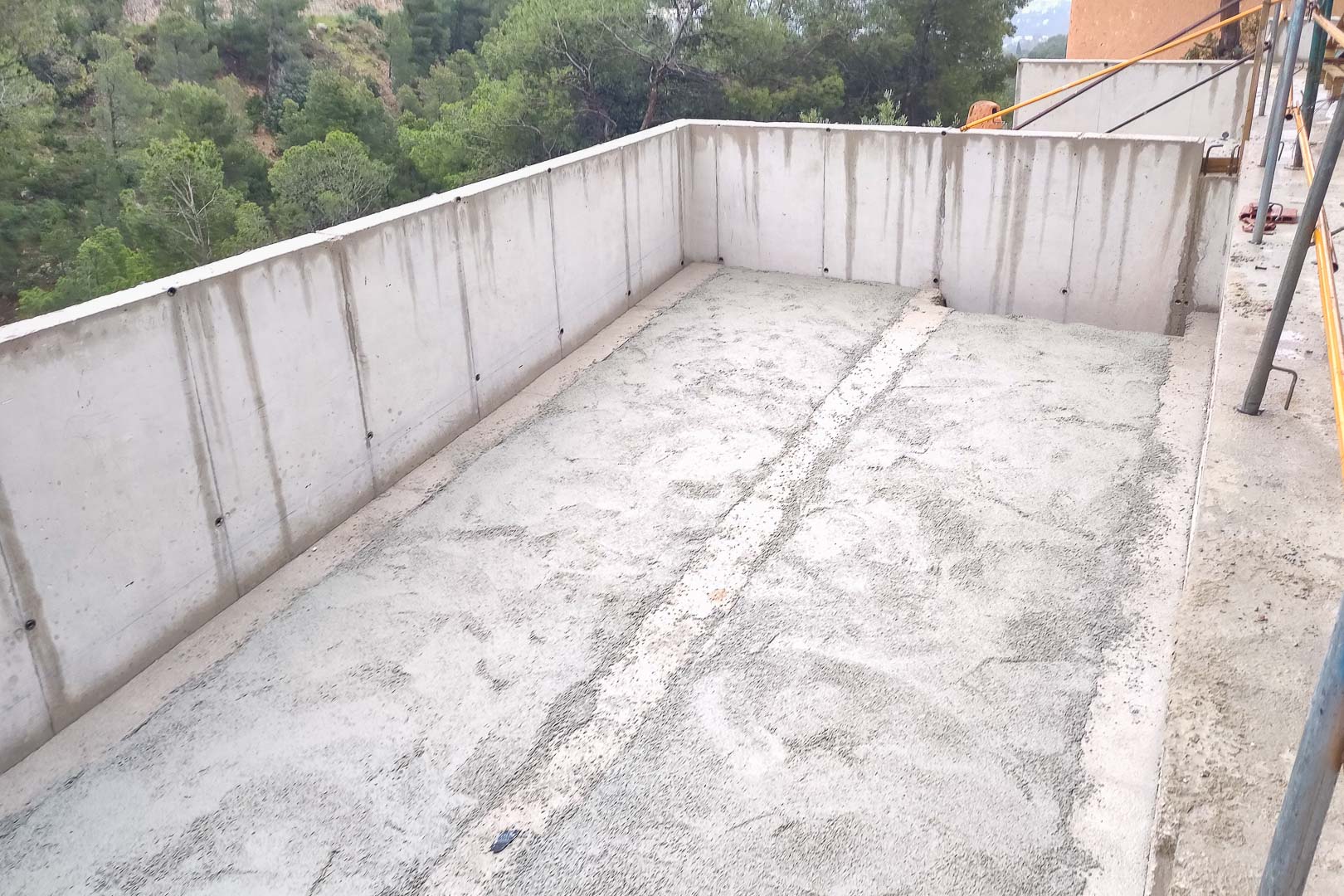Gewapende betonstructuur in de bouwfase, met de buitenmuren en vloerplaat van een toekomstig terras met uitzicht op het omliggende dennenbos.