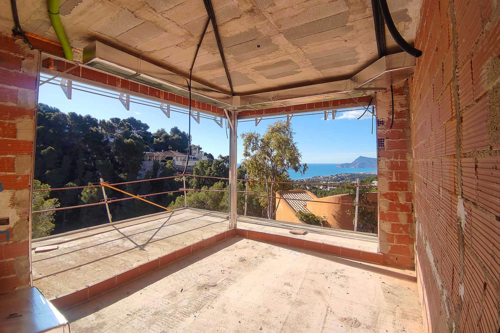 Panoramisch uitzicht op zee en de bergen vanuit een kamer in aanbouw, voorzien van grote hoekvensters en zichtbare technische leidingen.
