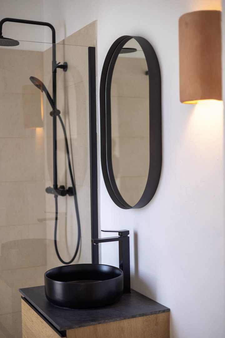 Badkamerdetail met een ronde zwarte wastafel, ovale spiegel en glazen douche.
