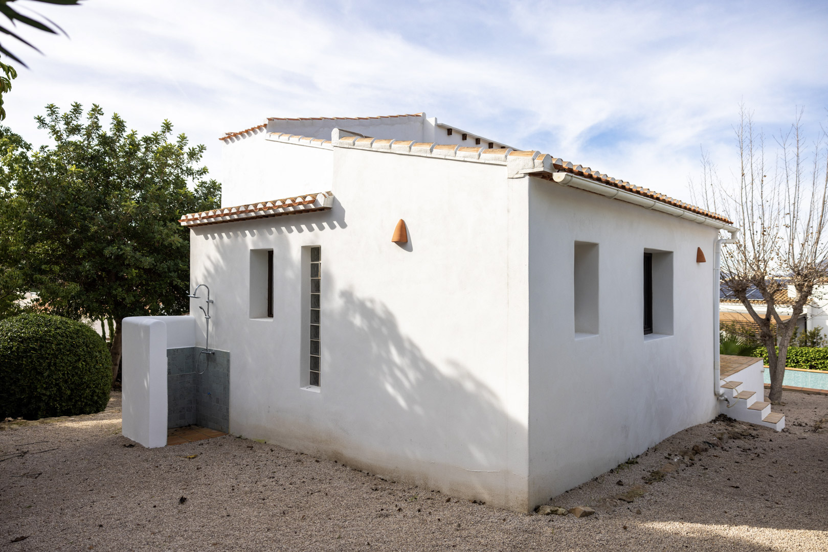 Buitenaanzicht van de villa met de betegelde buitendouche en minimalistische witte gevel met ramen.
