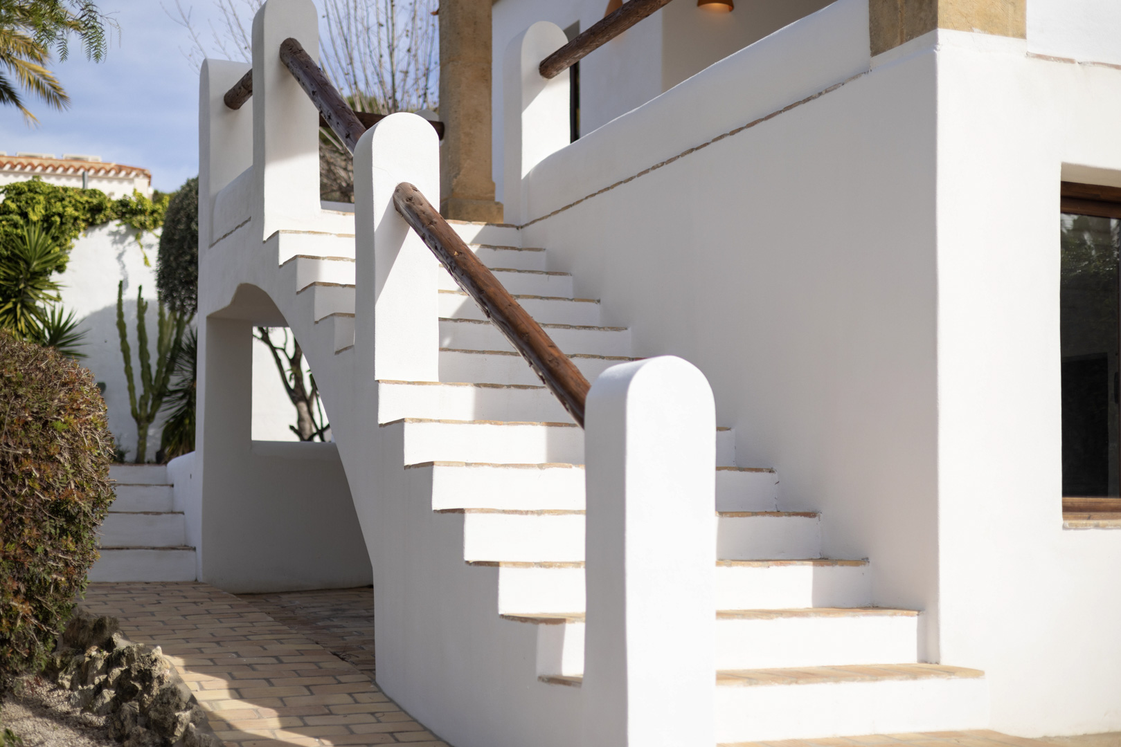 Detail van witte buitentrap met houten leuning en bakstenen bestrating.