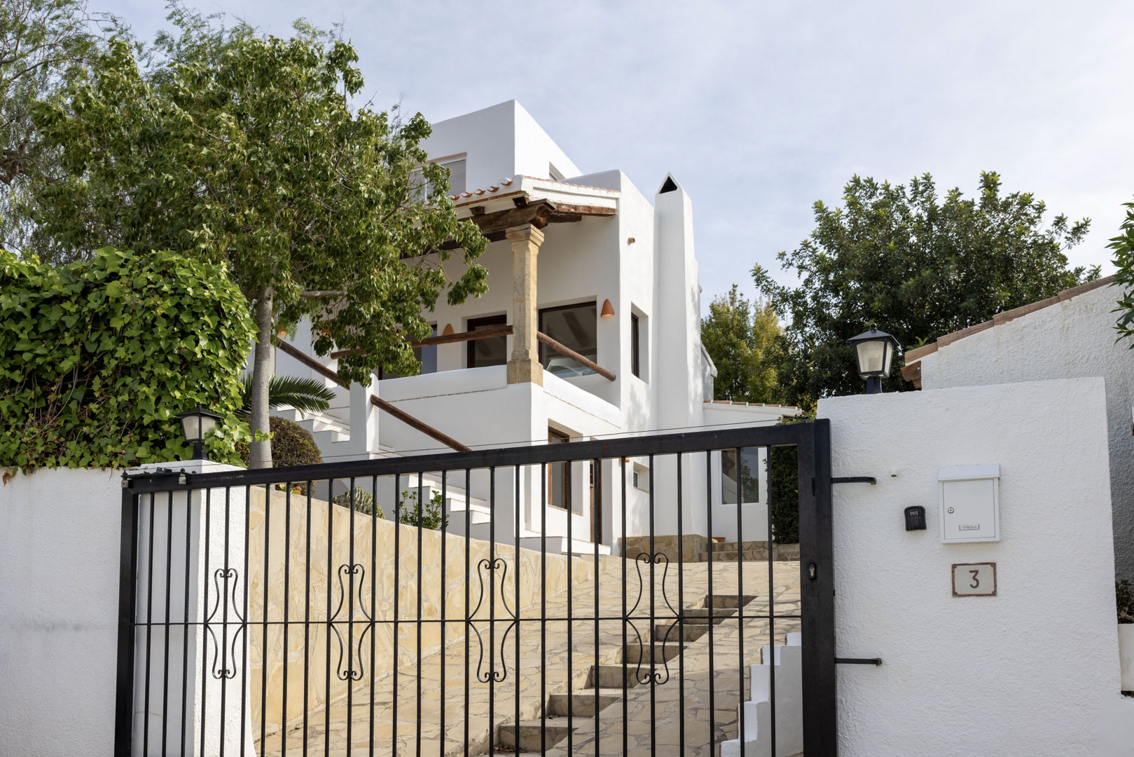 Exterieur van de villa met een smeedijzeren poort en witte gevel. Stenen details en weelderige vegetatie.