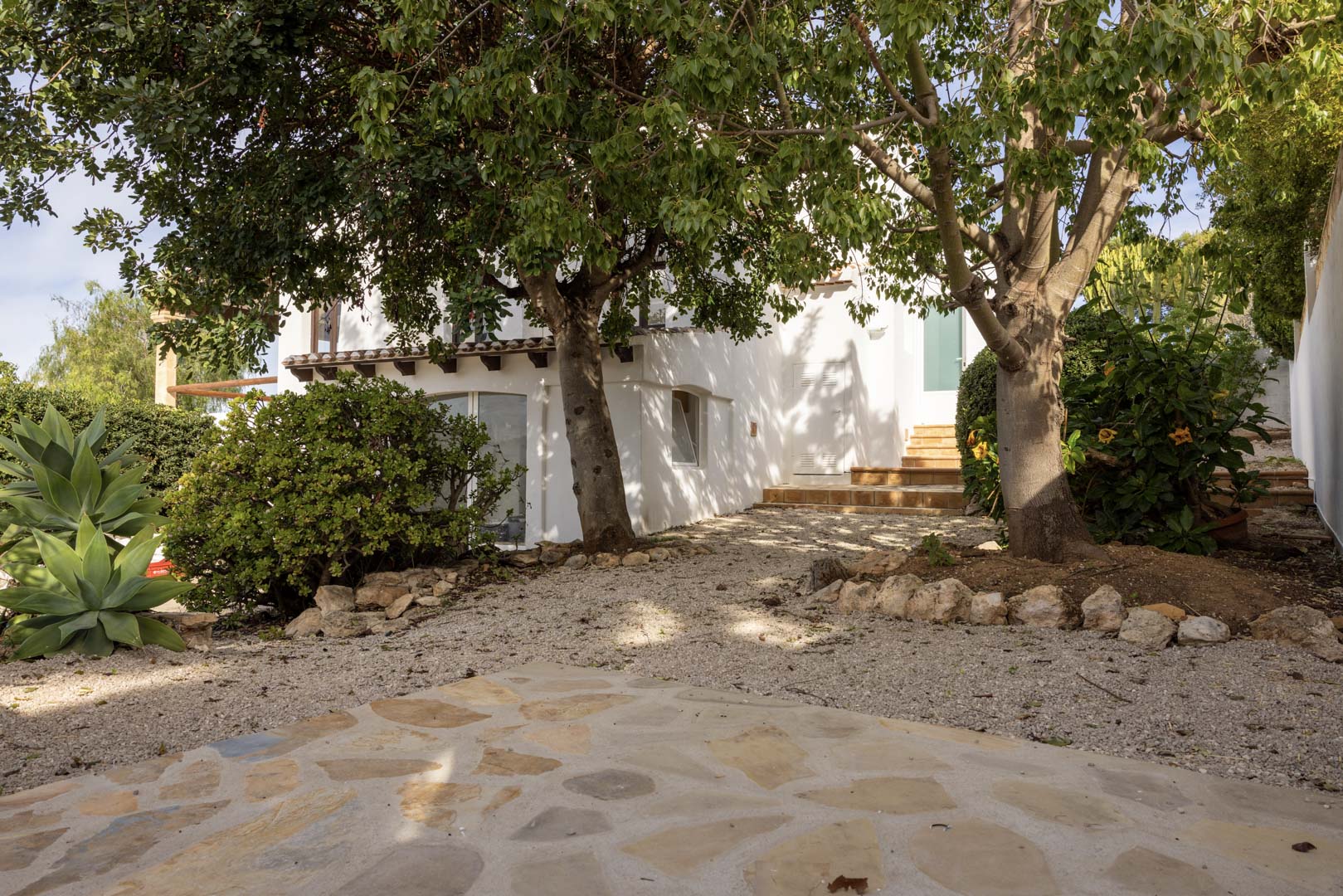 Exterieur van de villa met grindtuin en toegang tot de ingang. Volwassen groen en witte gevel.