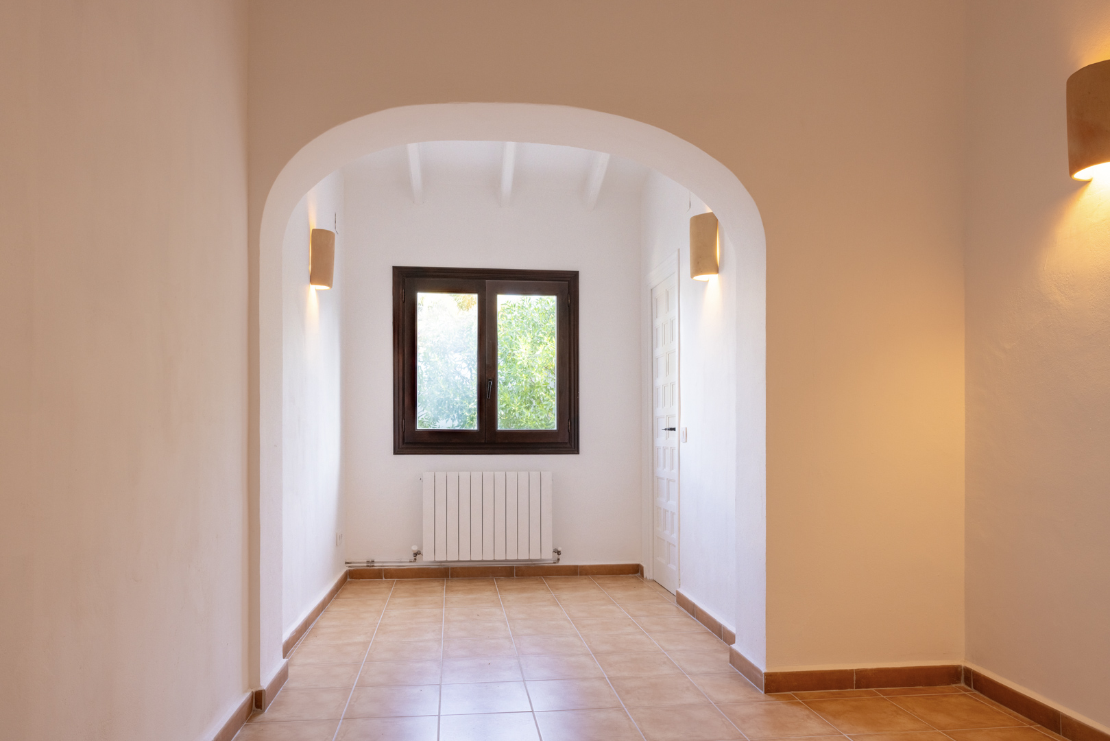 Licht interieur met een boogvormige ingang, keramische tegelvloer en een raam met donkere houten kozijnen.