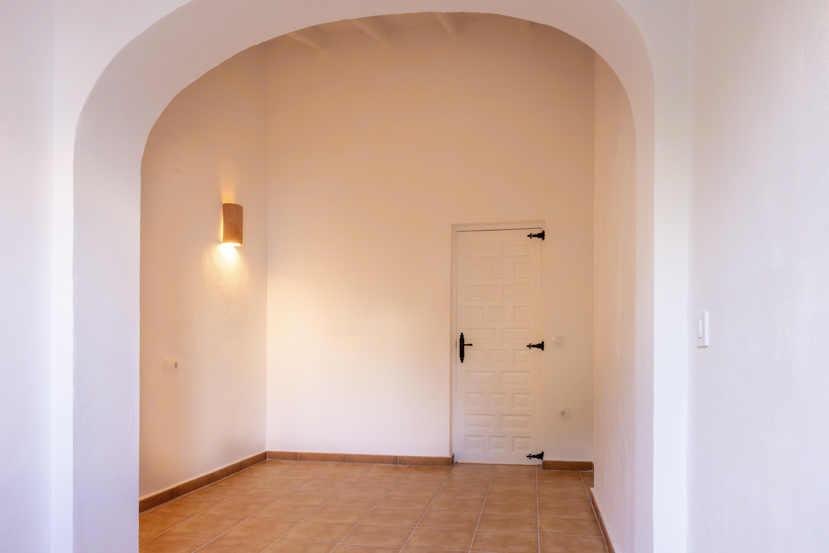 Lichte entree met boog, rustieke tegelvloer en witte deur met zwart beslag. Cilindervormige wandlamp.