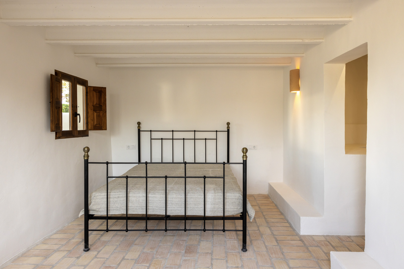 Lichte slaapkamer met smeedijzeren bed, houten luiken en rustieke tegelvloer.