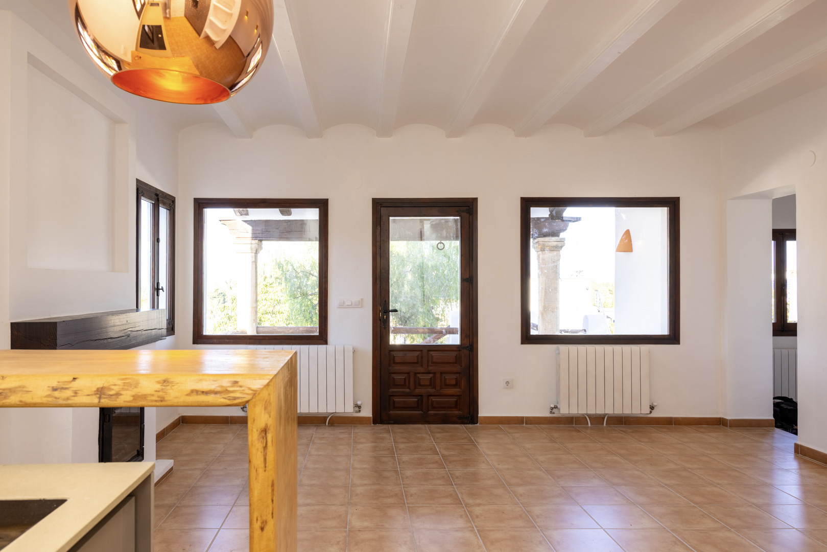 Lichte woonkamer met tegelvloer, balkenplafond en donkerhouten details. Uitzicht naar buiten.
