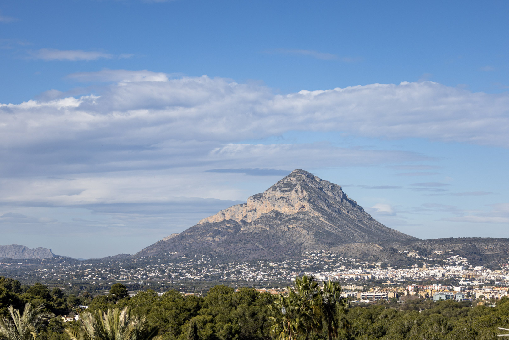 Panoramisch uitzicht op Montgó, Jávea, met een gedeeltelijk bewolkte hemel en vegetatie op de voorgrond.