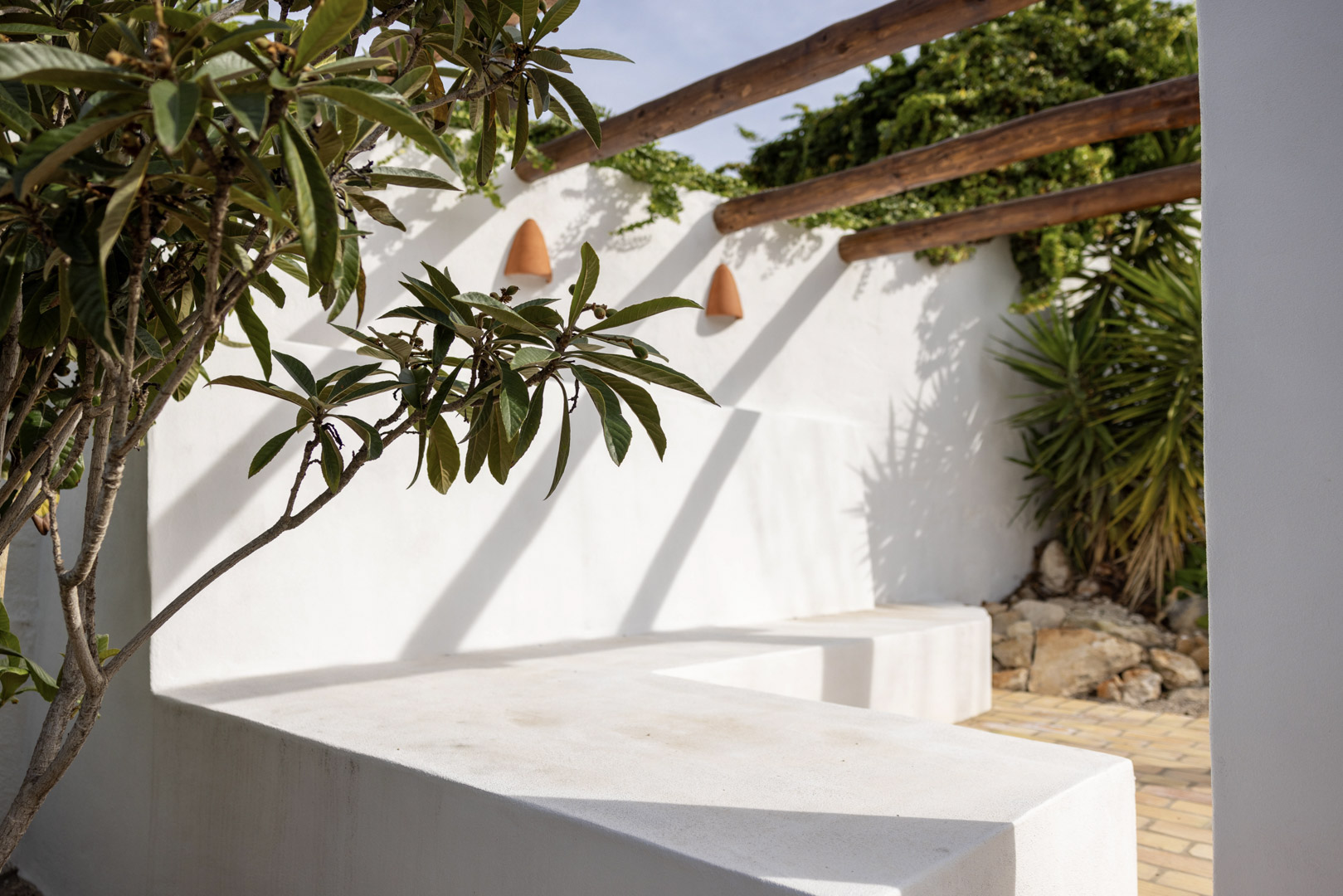 Terrasdetail met witgekalkte betonnen bank, houten pergola en weelderig groen.