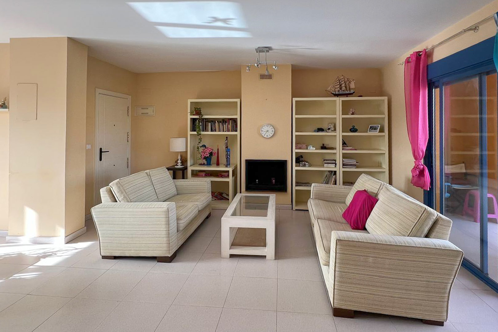Lichte woonkamer met gestreepte banken, witte stellingkasten en een decoratieve open haard. Lichte tegelvloer.
