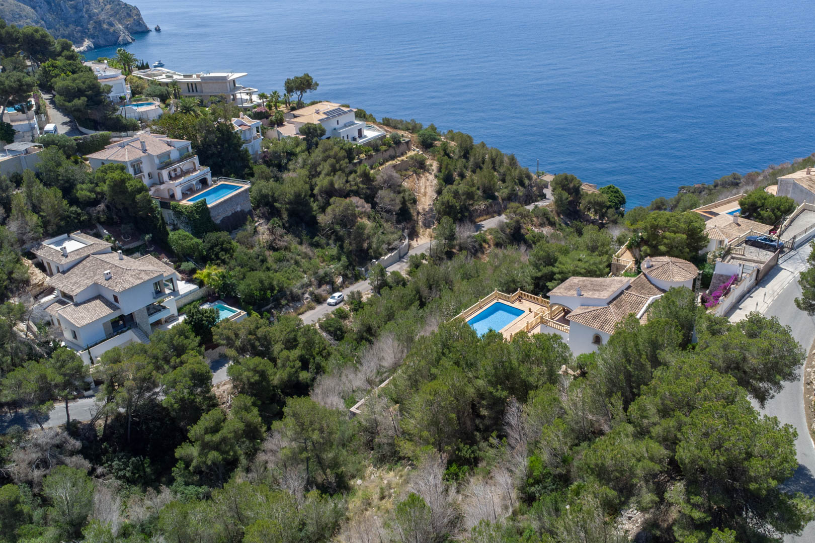 Luchtfoto van de kustlijn van Jávea met mediterrane villa's tussen de pijnbomen en de blauwe zee, wat de nabijheid van de percelen tot de kustlijn toont.