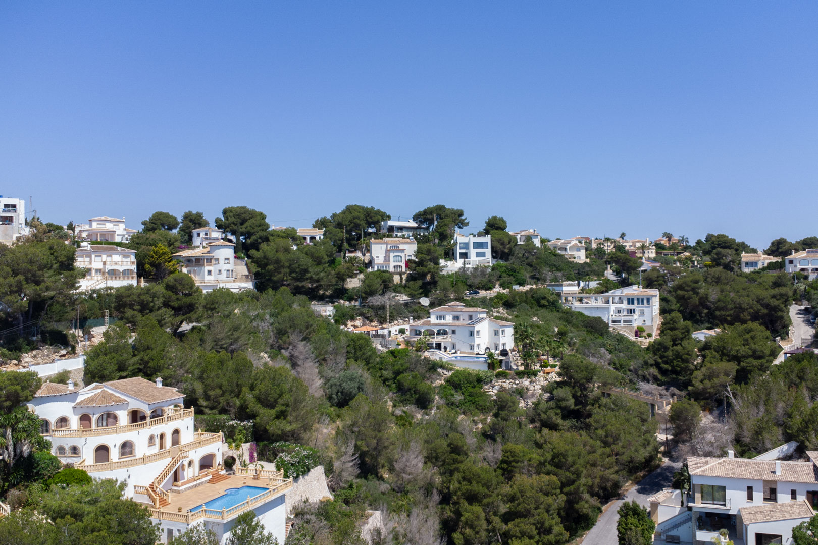 Verhoogd uitzicht op de woonwijk Costa Nova in Jávea, met mediterrane villa's en privézwembaden gelegen tegen een beboste heuvel met pijnbomen.