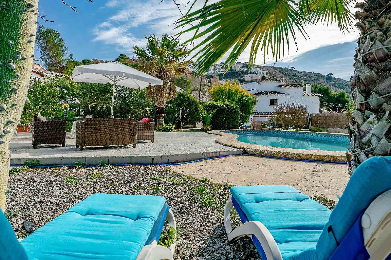 Buitenzwembad met niervormig bassin, stenen terras met rotan meubilair en blauwe ligbedden, omgeven door mediterrane beplanting en palmbomen.