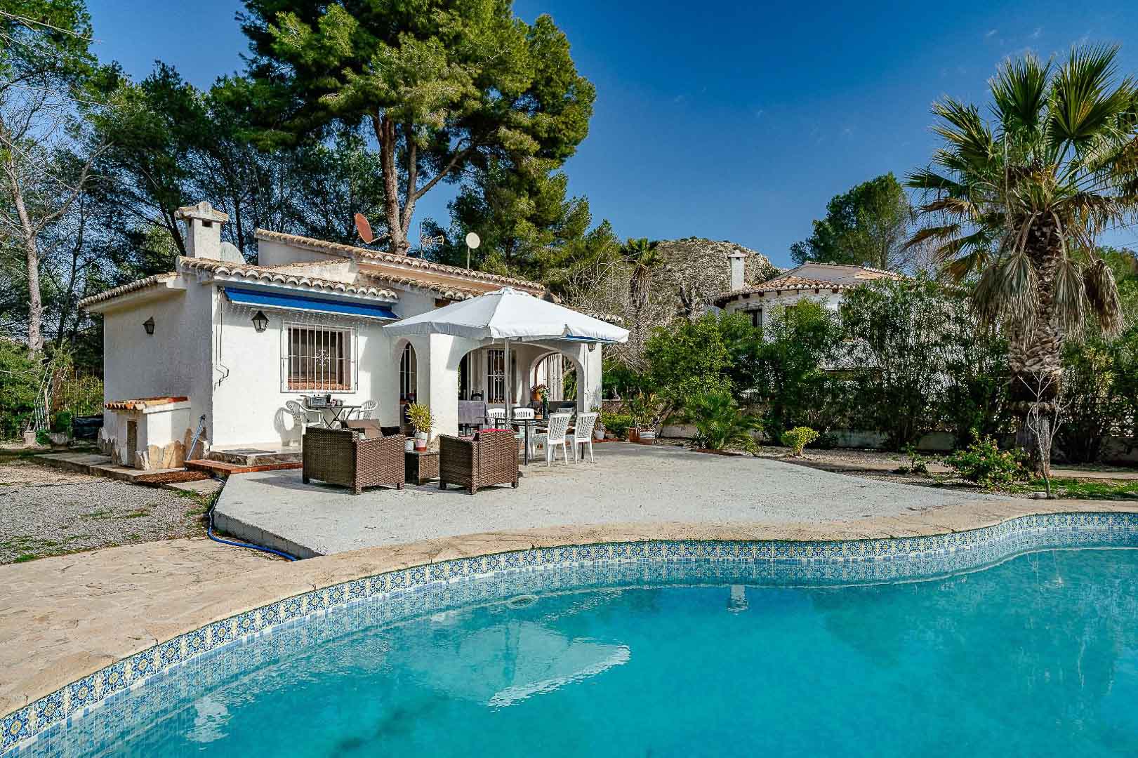 Exterieur van de villa met blauw mozaïekzwembad, ruim betegeld terras en een traditionele overdekte naya, omgeven door mediterrane pijnbomen.