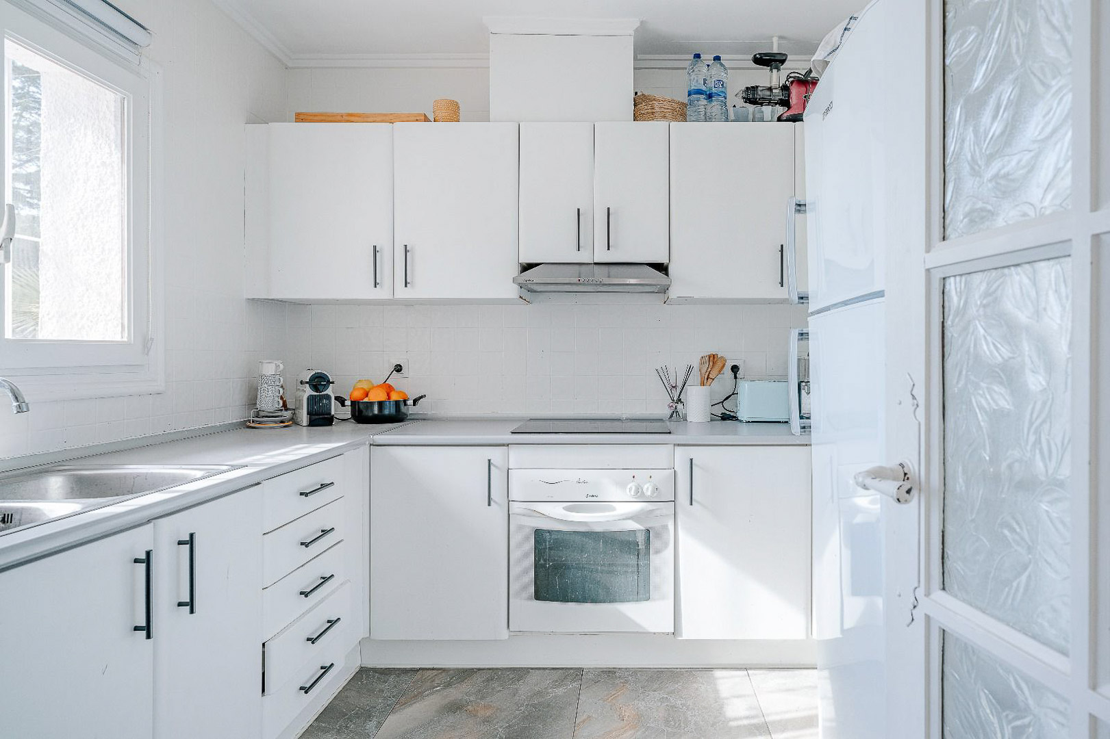 Functionele witte keuken met zwarte handgrepen, inbouwoven, keramische kookplaat en een dubbele spoelbak onder een raam met veel natuurlijk lichtinval.