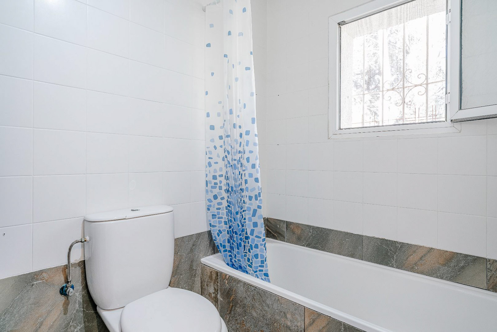 Gerenoveerde badkamer met een ingebouwd ligbad, wit keramisch toilet en lichte wandtegels. Een raam met beveiligingsrooster zorgt voor natuurlijk licht.