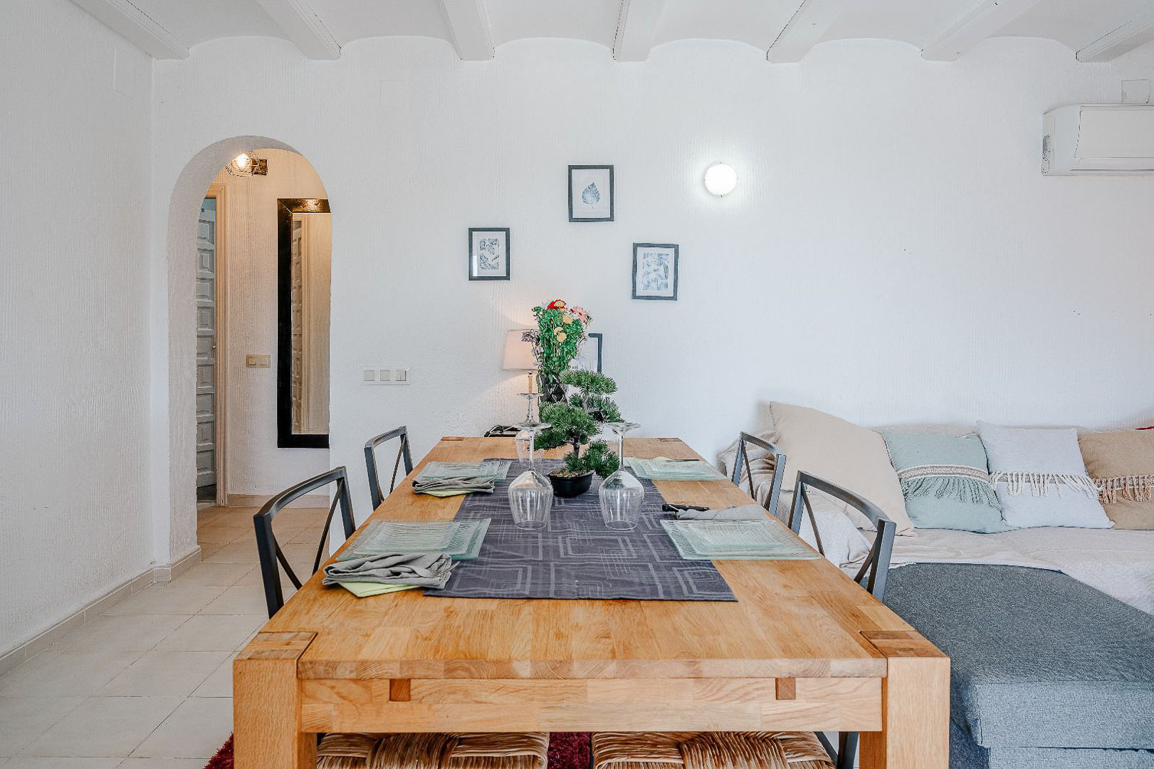 Lichte eethoek met een massief houten tafel en wit balkenplafond. Een ronde boog leidt naar de gang, grenzend aan de zithoek met bank en airconditioning.