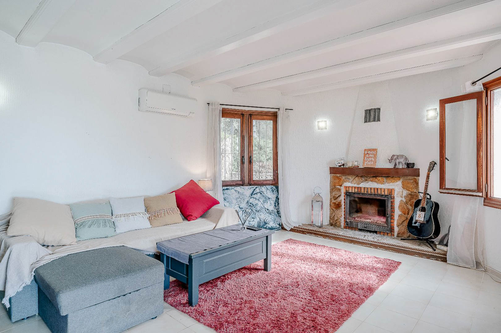 Lichte woonkamer met wit balkenplafond, een rustieke stenen open haard en airconditioning. Houten ramen laten veel natuurlijk licht binnen in deze mediterrane zithoek.