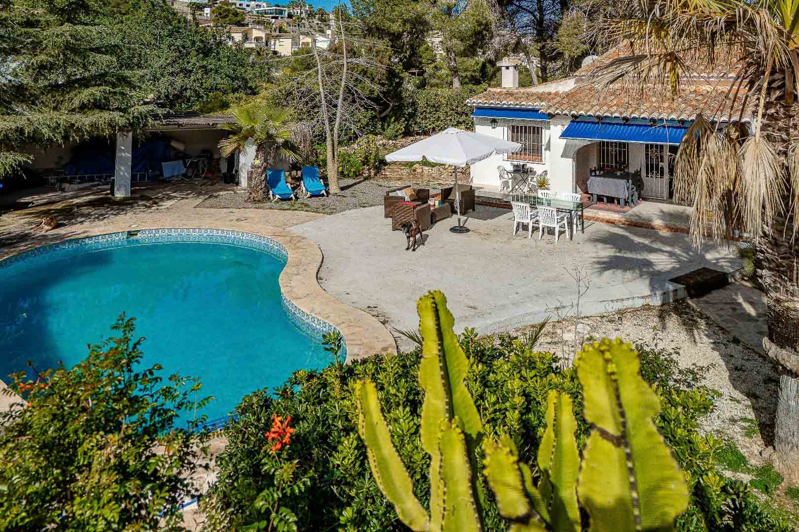Mediterrane buitenruimte met niervormig zwembad, ruim stenen terras met eethoek en een gelijkvloerse villa met karakteristieke blauwe zonneschermen.