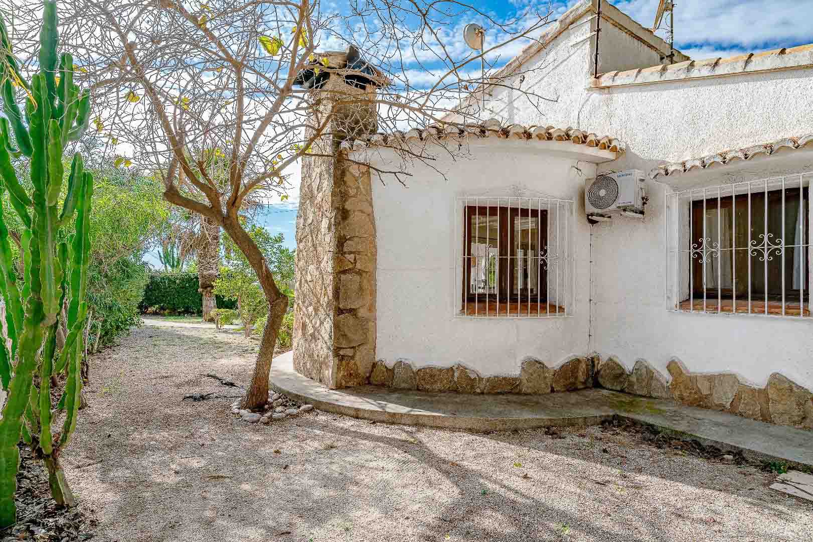 Mediterrane gevel met natuurstenen schoorsteen, houten kozijnen met wit siersmeedwerk en een onderhoudsvriendelijke tuin met grind en karakteristieke cactussen.