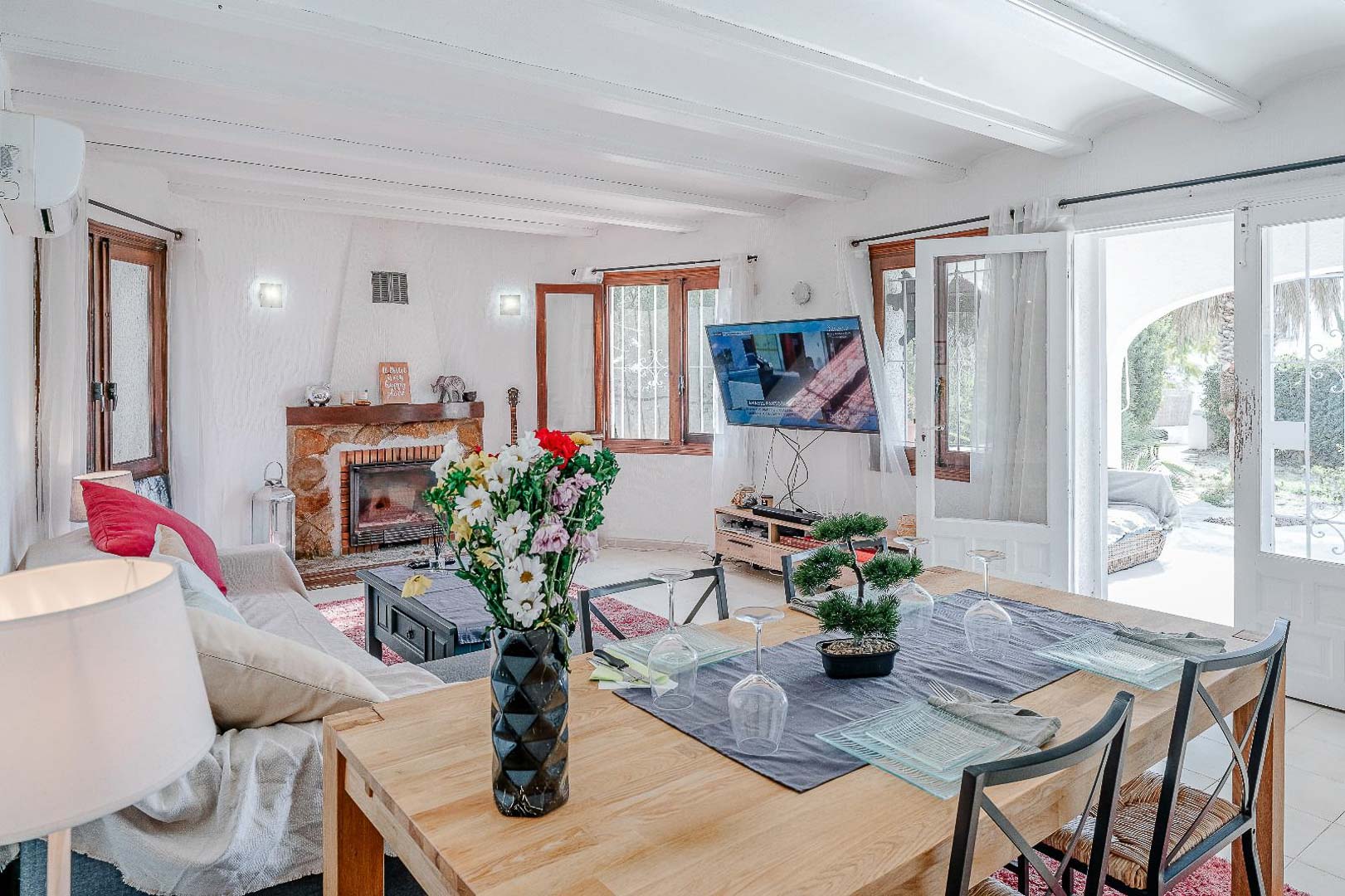 Open woon- en eetkamer met wit balkenplafond, een natuurstenen open haard en directe toegang tot het zonnige terras via openslaande deuren.
