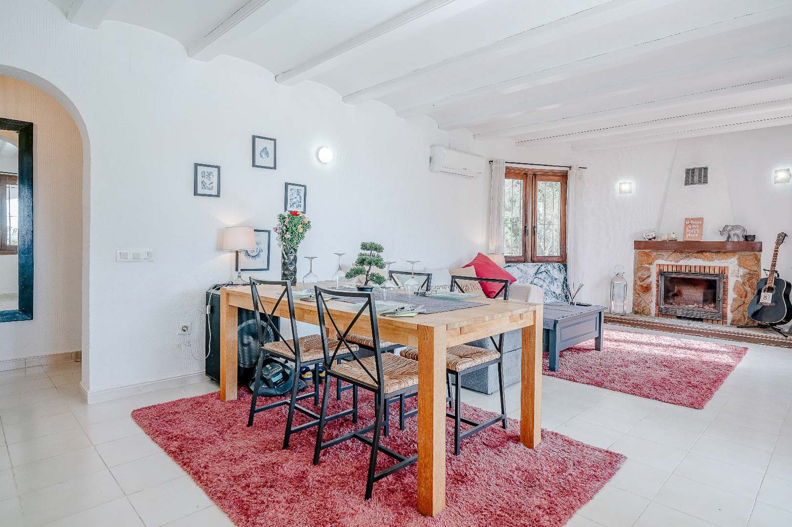 Open woon- en eetkamer met wit balkenplafond, een natuurstenen open haard en een houten eethoek geplaatst op rode decoratieve tapijten.