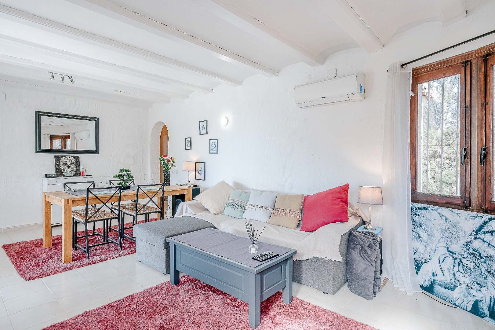 Open woon- en eetkamer met witgelakte balkenplafonds, split-airconditioning, lichte tegelvloeren en een houten eethoek in een mediterrane stijl.