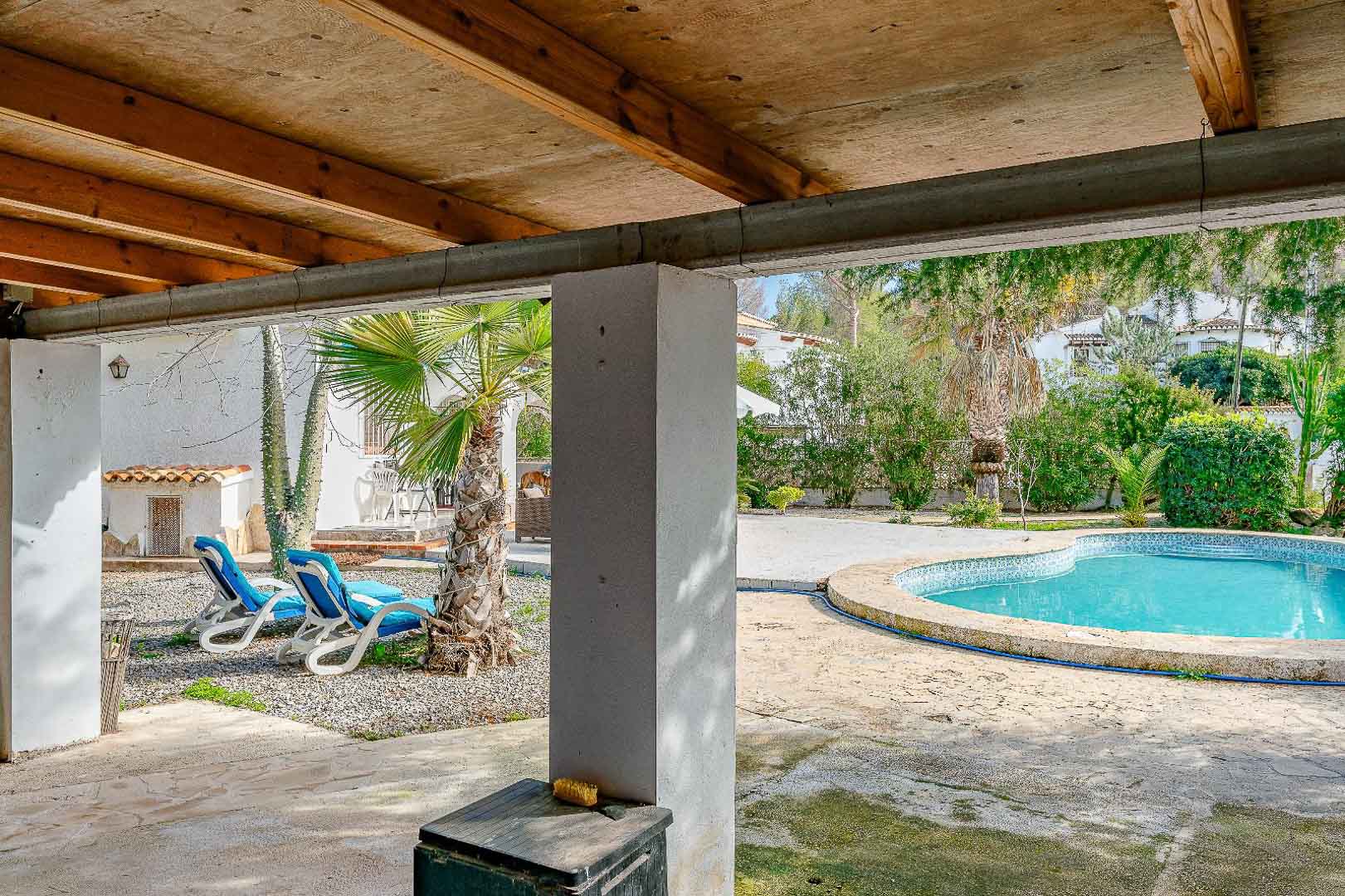 Overdekt terras met houten balkenplafond, uitkijkend op het niervormige zwembad en de mediterrane tuin met palmbomen en ligbedden.