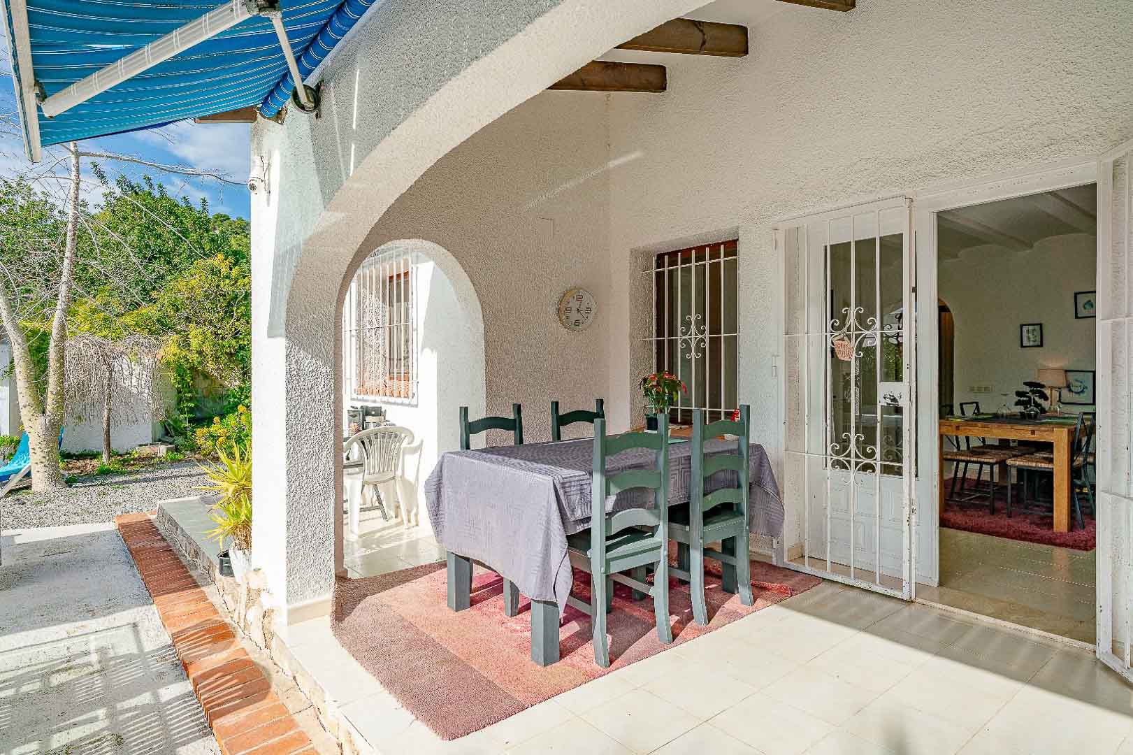 Overdekte veranda met mediterrane bogen, houten balken en een eethoek buiten. De ruimte verbindt de tuin met het interieur via beveiligde glazen deuren.