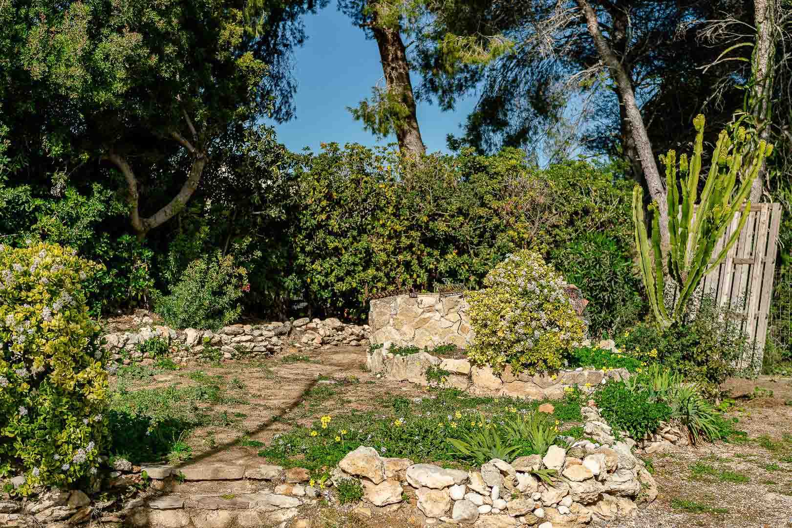 Rustieke tuin met drooggestapelde stenen muren, mediterrane beplanting en een centrale stenen put omgeven door pijnbomen en struiken.