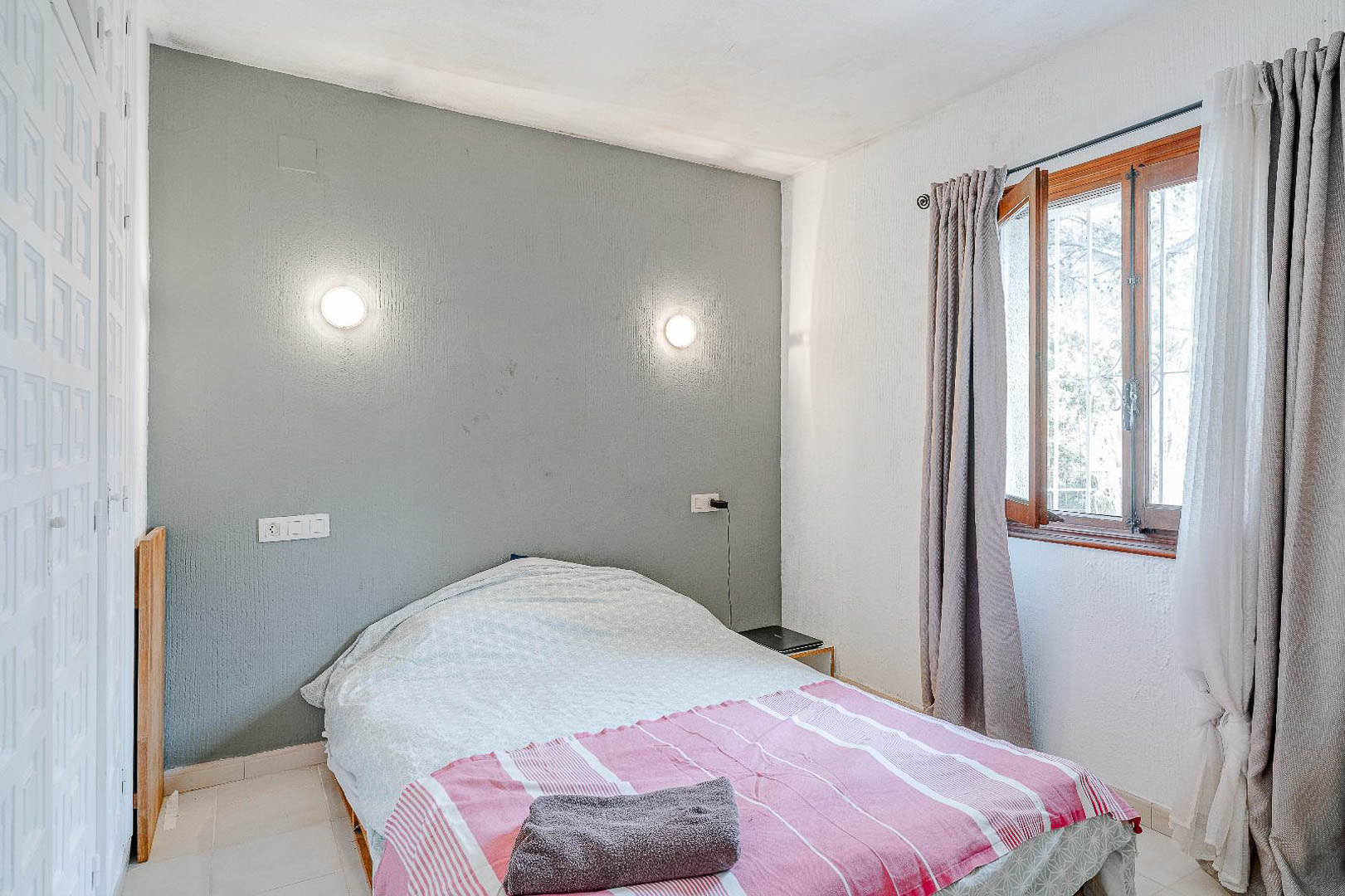 Slaapkamer met een grijze accentmuur, ronde wandlampen en een witte inbouwkast. Houten raamkozijn met grijze gordijnen biedt natuurlijke lichtinval.