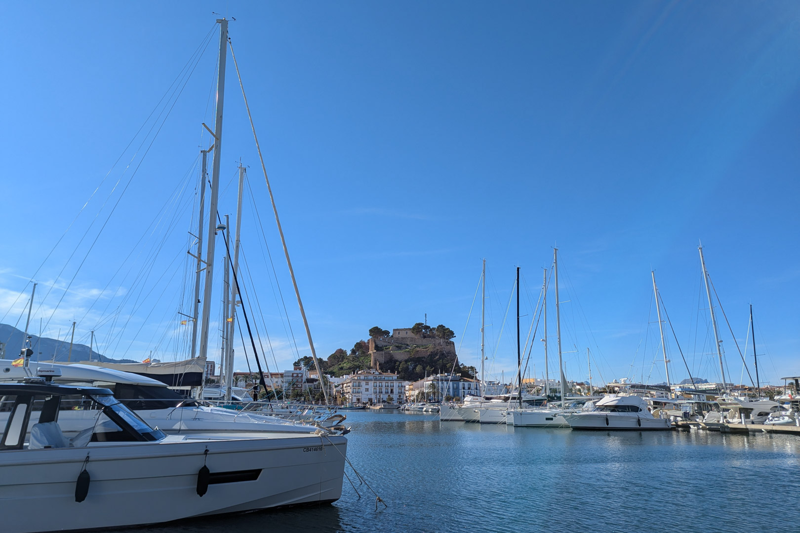 Uitzicht op de jachthaven met jachten en het kasteel van Dénia op de achtergrond onder een helderblauwe lucht.